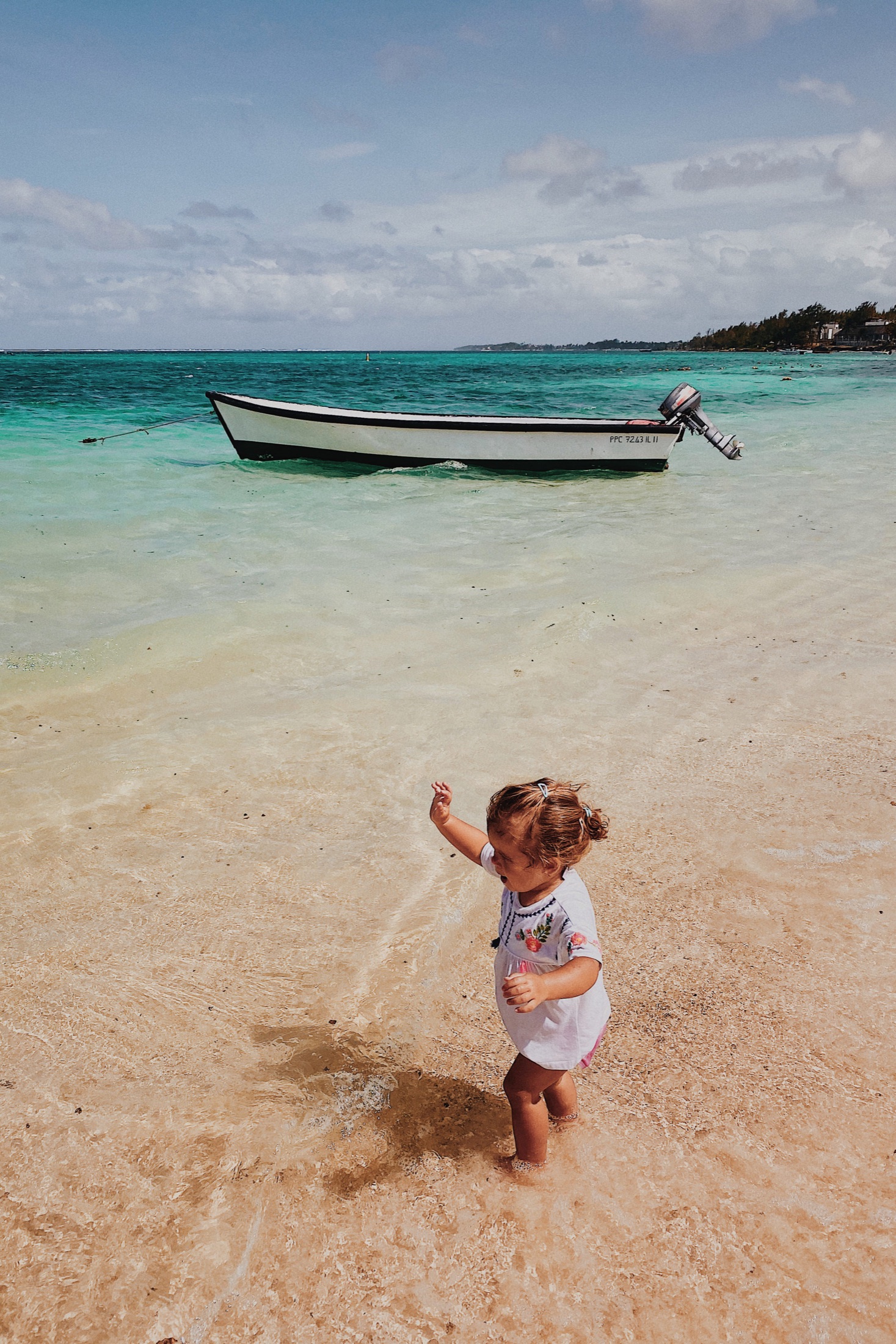 A child plays in shallow waves on a sandy beach, with a small boat beyond.